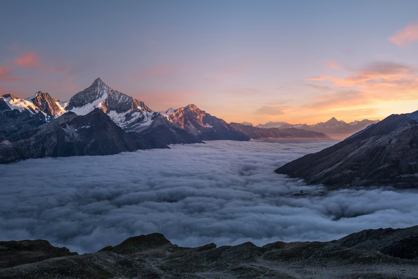 Snow-lit alpine peaks rising above a sea of clouds at dusk